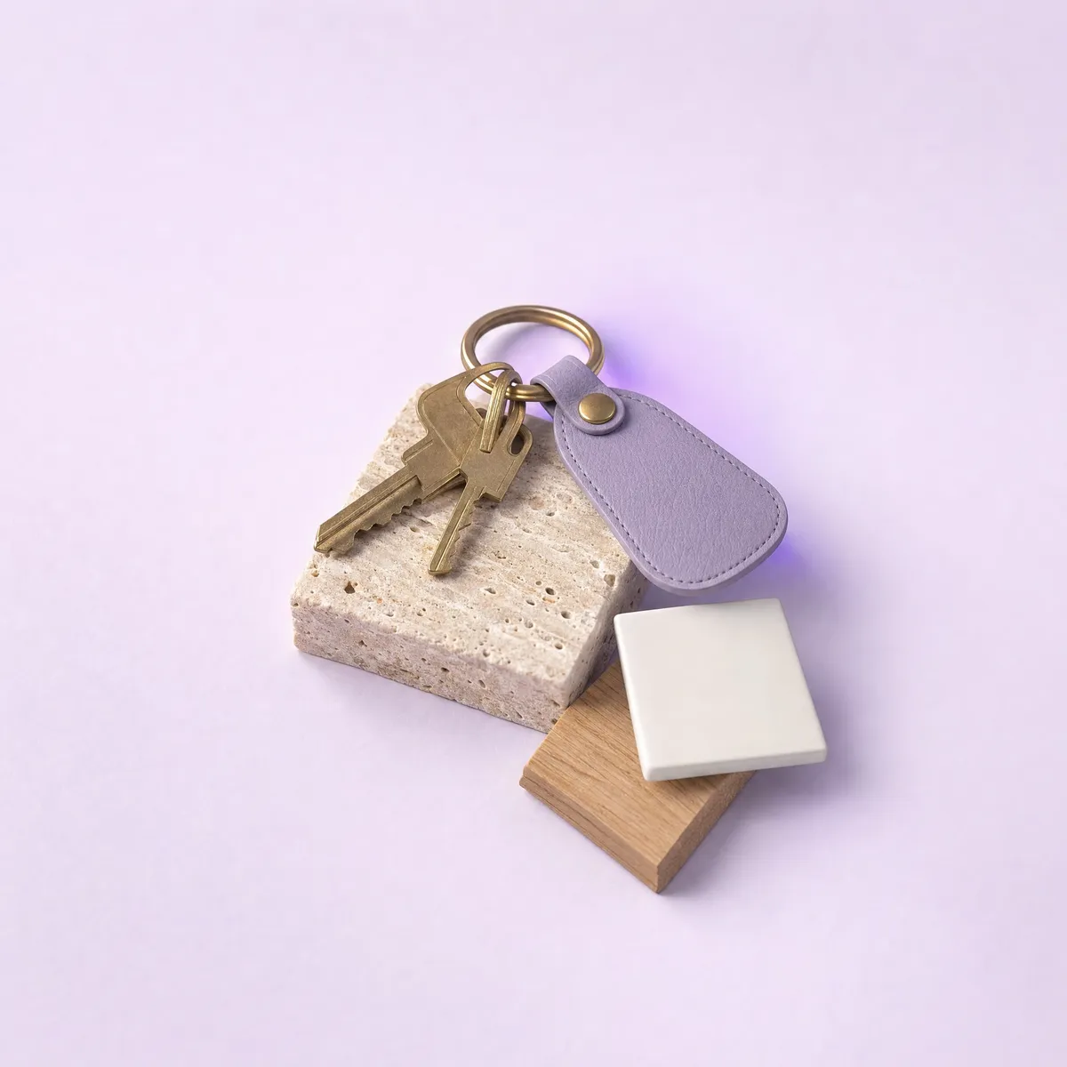 Brass house keys with a violet leather tag on a travertine block, beside a white tile and an oak offcut